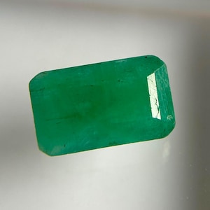 May include: A rectangular emerald gemstone with a vibrant green hue and a faceted surface. The stone displays natural internal inclusions and a polished finish, reflecting light on its top right corner, showcasing a clear, transparent quality.