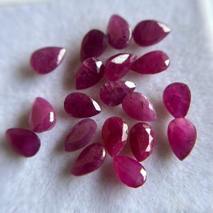 May include: A collection of fifteen loose, pear-shaped rubies with a faceted cut. The gemstones exhibit a deep, vibrant magenta-red color and are arranged in a cluster on a white, textured surface, showcasing their reflective surfaces.