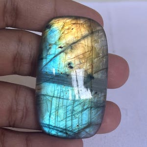 Pode incluir: Uma pedra preciosa de labradorita retangular polida. A pedra exibe um vibrante jogo de cores, com tons de azul, dourado e toques de verde. A gema é segurada em uma mão, mostrando sua beleza natural.