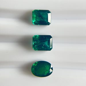 Pode incluir: Três pedras preciosas soltas de cor verde profunda com superfícies facetadas. Duas das pedras são retangulares com cantos cortados, enquanto a terceira pedra na parte inferior tem formato oval. Cada gema exibe inclusões internas e reflexos.