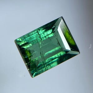 May include: A rectangular-cut gemstone with a deep forest green hue. The stone features a faceted surface that reflects light, showcasing internal inclusions and a transparent quality with varying shades of emerald and dark green throughout.