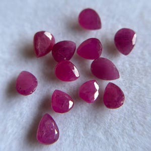 May include: A collection of eleven small, pear-shaped loose rubies. Each gemstone features a faceted surface that reflects light, displaying a deep, vibrant magenta-red color throughout the stones, arranged in a scattered formation.