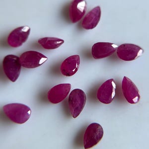 Puede incluir: Una colección de pequeños rubíes sueltos en forma de pera con un tono rojo magenta intenso. Cada piedra preciosa presenta una superficie facetada que refleja la luz, mostrando una silueta de lágrima dispuesta en un patrón disperso.