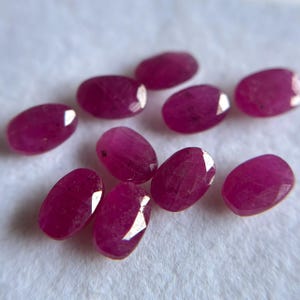 May include: A collection of nine oval-shaped, faceted rubies with a deep magenta-red color. Each gemstone features a polished surface with visible internal inclusions and light-reflecting facets arranged in a scattered formation.
