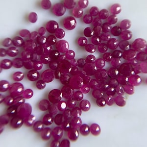May include: A collection of numerous small, round, faceted rubies in varying shades of deep pink and red. Each gemstone features a polished surface with multiple triangular facets that reflect light, creating a sparkling appearance across the group.