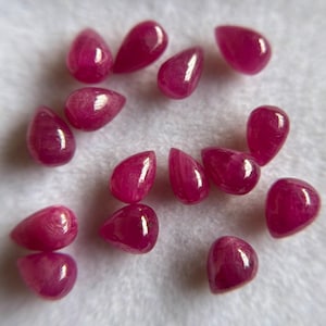 May include: A collection of fourteen loose, teardrop-shaped rubies with a vibrant magenta-red hue. Each gemstone features a smooth, polished surface with subtle internal inclusions and light reflections, arranged in a scattered pattern.