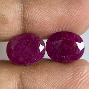 Puede incluir: Dos rubíes facetados de forma ovalada con un color rojo magenta profundo y saturado. Cada piedra preciosa presenta una superficie pulida con múltiples facetas geométricas que reflejan la luz, mostrando una calidad translúcida y una estructura cristalina interna distintiva.