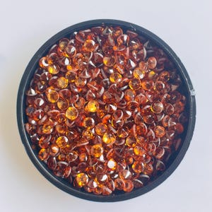 May include: A black container filled with numerous small, round, faceted gemstones. The gemstones are a warm amber color, with variations in shade from light orange to deep reddish-brown. The gems are densely packed, filling the container.