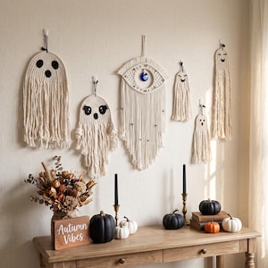 May include: A collection of off-white macrame wall hangings featuring ghost shapes with black felt eyes and mouths, and a central eye-shaped piece with a blue glass bead. The set includes six distinct fiber art pieces suspended on a wall.