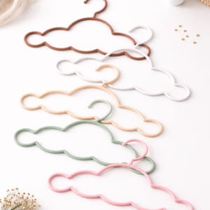 May include: A collection of cloud-shaped clothes hangers in various pastel colors, including brown, white, beige, green, and pink. The hangers are arranged on a white surface, suggesting a nursery or children's room theme.