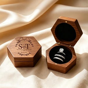 Puede incluir: Dos joyeros hexagonales de madera, uno cerrado y otro abierto. La caja cerrada tiene pájaros e iniciales grabadas. La caja abierta muestra dos anillos con piedras claras, sobre un interior de terciopelo negro. Las cajas son de madera oscura.