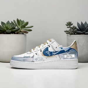 May include: White and blue Nike Air Force 1 sneakers with a unique design. The shoe features a white base with blue and tan accents, including a blue swoosh and a patterned design on the side. The sole is white, and the laces are tan.