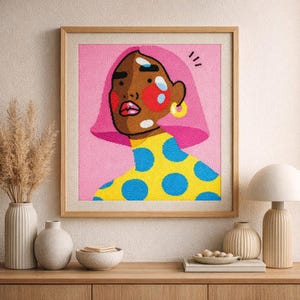May include: Framed artwork featuring a stylized portrait of a person with brown skin, pink hair, and yellow earrings. The person wears a yellow top with blue polka dots against a pink background. The artwork is in a wooden frame.