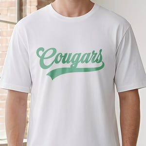 Peut inclure: T-shirt blanc avec le mot "Cougars" en vert en écriture cursive. Le mot est légèrement incliné et souligné en vert. Le t-shirt a des manches courtes et un col rond.