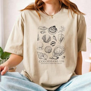 May include: A beige short-sleeved t-shirt featuring a black line-art illustration of various seashells arranged in a cluster. Below the shells, the text reads COLOSSIANS 1:16 FOR BY HIM ALL THINGS WERE CREATED in a clean, serif font.
