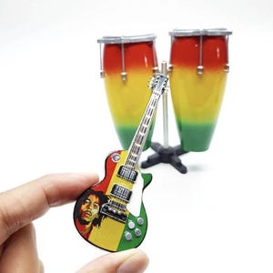 May include: A miniature electric guitar pin featuring a portrait of a man with dreadlocks and a red, yellow, and green body, positioned in front of a pair of red, yellow, and green conga drums mounted on a black stand with a silver metal pole.