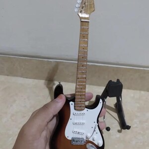Puede incluir: Miniatura de guitarra el&eacute;ctrica con acabado sunburst, golpeador blanco y herrajes cromados. La peque&ntilde;a guitarra es marr&oacute;n, blanca y negra. Est&aacute; sobre un soporte negro.