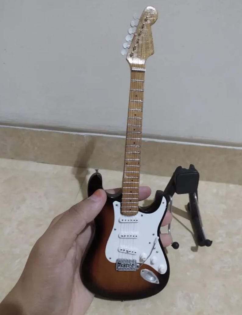 Puede incluir: Miniatura de guitarra el&eacute;ctrica con acabado sunburst, golpeador blanco y herrajes cromados. La peque&ntilde;a guitarra es marr&oacute;n, blanca y negra. Est&aacute; sobre un soporte negro.