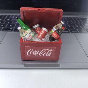 Op de afbeelding: Een kleine rode Coca-Cola-koeler gevuld met ijs en diverse miniatuurflessen en -blikjes. De koeler heeft het Coca-Cola-logo op de voorkant. De koeler is open en toont de inhoud.