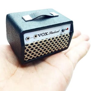 May include: A miniature black guitar amplifier with a textured exterior, a top carrying handle, and a white front control panel featuring the text VOX Student. The speaker area is covered by a beige mesh grille with a hexagonal pattern.
