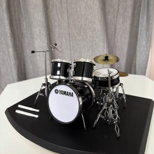 May include: Miniature black and silver Yamaha drum set with a white bass drum head. The set includes a snare drum, two toms, a cymbal, and drumsticks, all on a black base.