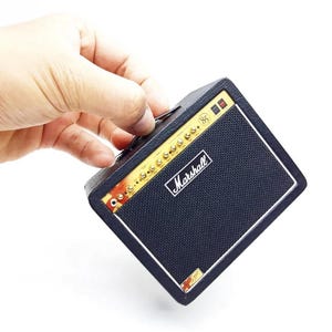 May include: A black Marshall guitar amplifier with a textured finish, a gold-colored control panel featuring multiple knobs, a white Marshall logo on the black speaker grille, and a small gold emblem with the number 85 on the top right corner.