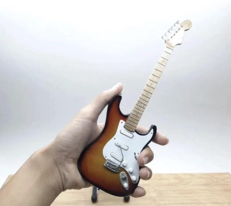 Puede incluir: Miniatura de guitarra el&eacute;ctrica con acabado sunburst, golpeador blanco y herrajes cromados. La guitarra tiene m&aacute;stil y pala de madera. El instrumento es una peque&ntilde;a r&eacute;plica de una guitarra el&eacute;ctrica de tama&ntilde;o completo.