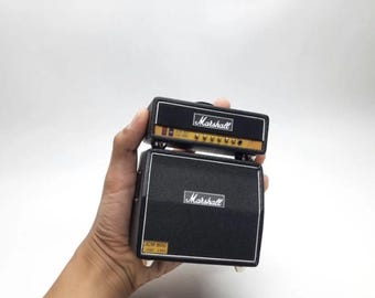 Può includere: Un amplificatore per chitarra Marshall nero in miniatura. La testata dell'amplificatore superiore ha un pannello di controllo dorato e il logo Marshall. Il cabinet inferiore presenta il logo Marshall e la scritta "JCM 800 LEAD 1960".