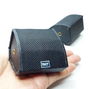 May include: Two small black wedge-shaped speakers with a textured mesh front grille. Each speaker features a gold-coloured circular input port on the side and a small rectangular blue and white logo plate on the front that reads TACT PRO-AUDIO.