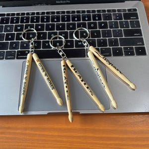 Op de afbeelding: Sleutelhangers met miniatuur drumstokken met de namen "GUNS 'N' ROSES" en "BEATLES" in zwart. De drumstokken zijn gemaakt van lichtgekleurd hout en bevestigd aan zilveren sleutelringen. De sleutelhangers worden weergegeven op een laptoptoetsenbord.