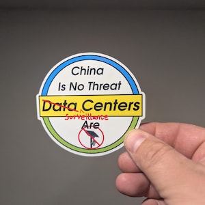May include: A circular sticker with a white background, blue and green borders, and black text reading China Is No Threat. The words Data Centers are crossed out in red, with the word Surveillance written above and Are below a red prohibition symbol.