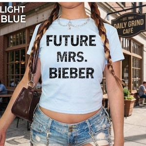 May include: Light blue t-shirt with the text "FUTURE MRS. BIEBER" in black. The shirt is paired with denim shorts. A brown purse hangs from a shoulder strap. The background includes a cafe sign and outdoor seating.