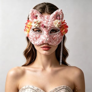 May include: A pink cat-shaped mask covered in small fabric roses, featuring rhinestone-lined eye openings, decorative cream and orange fabric flowers on the sides, and a long, dangling strand of clear beads hanging from the right side of the mask.