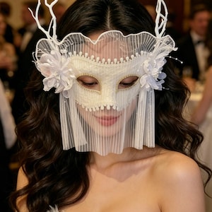 May include: A white masquerade mask with a delicate veil, floral accents, and a decorative antler-like headpiece. The mask has gold detailing around the eyes and is designed for formal events.