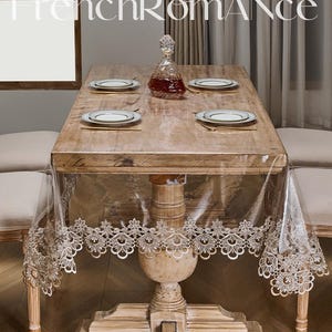 May include: A wooden dining table set for four, with a clear tablecloth featuring a decorative lace trim. The table is set with white plates with gold rims, a crystal decanter, and the text "French Romance" at the top.