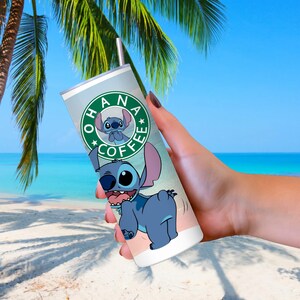 Peut inclure: Un gobelet en acier inoxydable avec un motif de personnage de dessin anim&eacute;. Le gobelet a un fond bleu clair avec les mots "OHANA COFFEE" dans un logo circulaire. Le personnage est bleu avec de grandes oreilles et une expression espi&egrave;gle.