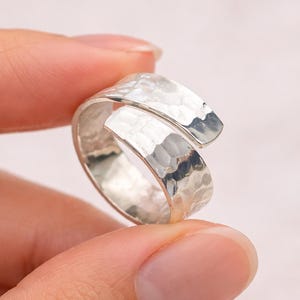 May include: A silver wrap ring with a hammered finish. The ring's design is open, with overlapping ends. The ring is held by a person's fingers against a soft pink backdrop.