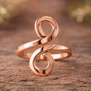 Peut inclure: Bague en or rose au design tourbillonnant. La bague présente deux éléments circulaires reliés par une bande incurvée. Le métal est poli et réfléchit la lumière. L'arrière-plan est flou, avec des tons naturels. Bijou.