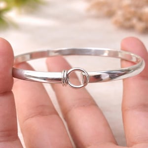 Puede incluir: Un brazalete de plata brillante con un diseño sencillo y elegante. El brazalete presenta un cierre circular con un pequeño cierre de barra. La superficie lisa y pulida refleja la luz, resaltando su estilo minimalista.