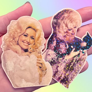 May include: Two die-cut stickers featuring a portrait of a blonde person with voluminous hair holding a small kitten. One sticker has a matte finish with warm tones, while the other features a holographic overlay with a repeating dot pattern.