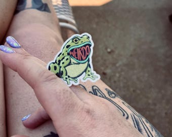 May include: A die-cut sticker featuring a green frog with dark spots and a wide-open mouth. Inside the frog's mouth, the word WEIRDO is printed in bold red capital letters. The sticker has a white border and a matte finish, showcasing a detailed illustration.