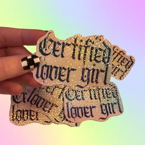 May include: A collection of die-cut stickers with a holographic glitter finish. Each sticker features the text "Certified lover girl" written in a bold, black gothic-style font. The stickers have an irregular, wavy edge shape and a reflective surface.