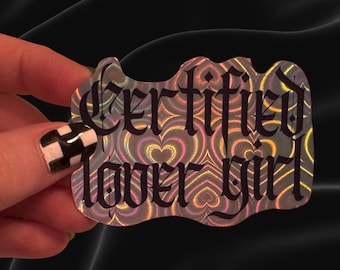 May include: A die-cut sticker with a holographic background featuring a repeating pattern of hearts in shades of pink, yellow, and blue. The text "Certified lover girl" is printed across the center in a bold, black, gothic-style font.