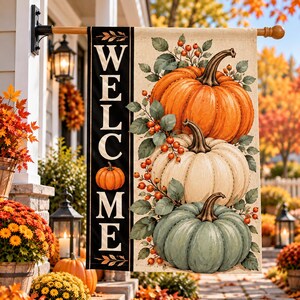 May include: A vertical garden flag featuring the word WELCOME in white serif letters on a black side panel. The main area displays three stacked pumpkins in orange, cream, and sage green, accented with green leaves and small orange berries.