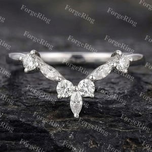 May include: A curved, silver-toned ring with multiple clear gemstones. The ring features a series of round and marquise-cut stones set in a delicate, symmetrical design. The ring is displayed against a dark, textured background.