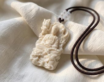 Può includere: Un ciondolo rettangolare color crema raffigurante una figura scolpita di Buddha seduto circondato da un motivo a drago. Il ciondolo è fissato a una collana con cordino marrone scuro, impreziosita da due piccole perline bianche traslucide.