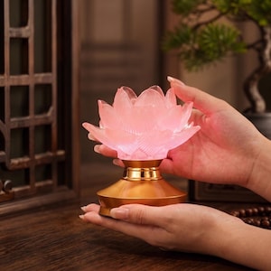 May include: A glowing pink lotus flower lamp featuring translucent, textured petals that emit a soft light. The flower rests upon a smooth, metallic gold base with a wide, circular bottom, providing a stable foundation for the decorative light fixture.