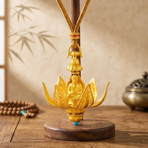Könnte beinhalten: Eine goldfarbene Buddha-Figur, die in einer offenen Lotusblüte sitzt und an einem dunklen Holzständer an einer geflochtenen Kordel hängt. Die Lotusblätter haben komplizierte Ausschnittmuster und der Sockel ist ein rundes Holzstück mit einer blauen Perle.