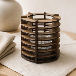 Op de afbeelding: Een cilindrische kaarshouder gemaakt van donkerbruin gebeitste houten latten in een verticaal, open frameontwerp. Binnenin de houten structuur bevindt zich een kleine, doorzichtige glazen votiefhouder met verticale ribbels aan de buitenkant.