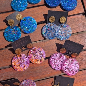 Puede incluir: Una colección de pendientes redondos de purpurina hechos a mano en varios colores, incluyendo azul, morado y rosa. Cada par tiene un perno de color dorado y se muestra en una tarjeta negra para pendientes. Sobre una superficie de madera.